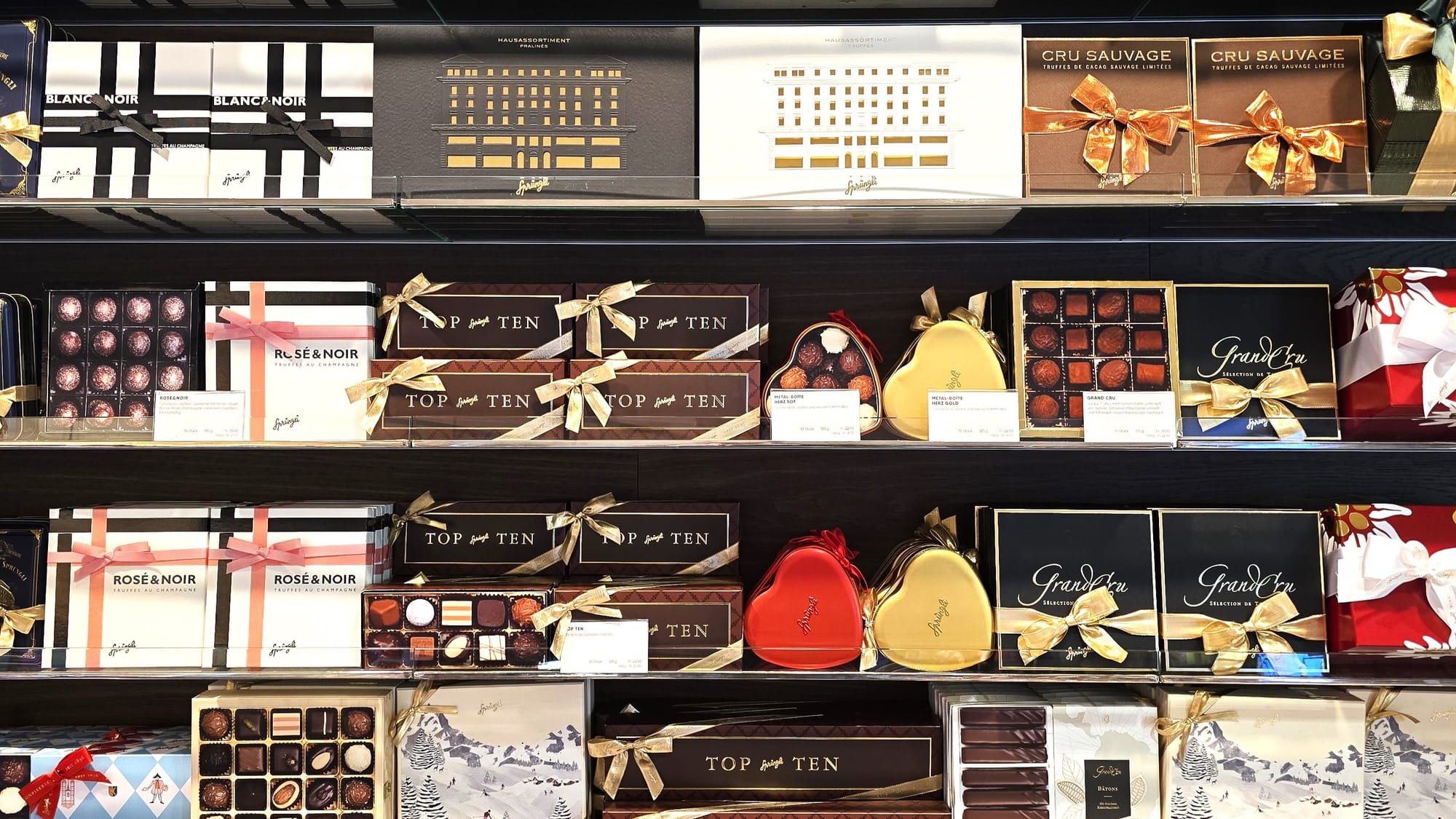 Edle Pralinen-Geschenkboxen von Sprüngli, dekorativ verpackt mit Schleifen, in verschiedenen Varianten und Größen auf Regalen präsentiert – auch im Online-Shop erhältlich.